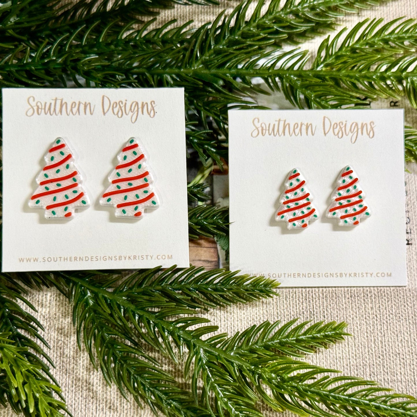 Pair of Christmas tree earrings on 'Southern Designs' cards with green foliage in the background.
