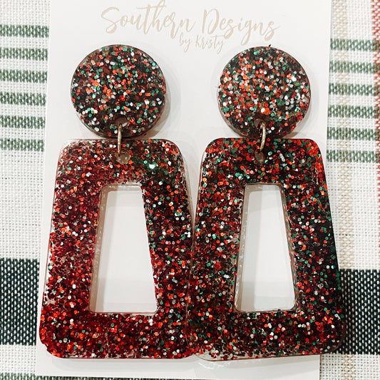 A pair of glittery red and black Christmas-themed dangle earrings displayed on a textured fabric background with 'Southern Designs' branding.