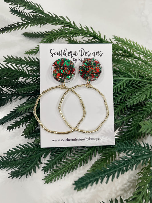 A pair of dangle earrings with red and green circular designs, displayed on a white card with 'Southern Designs by Kristy' printed on it, surrounded by green pine branches.