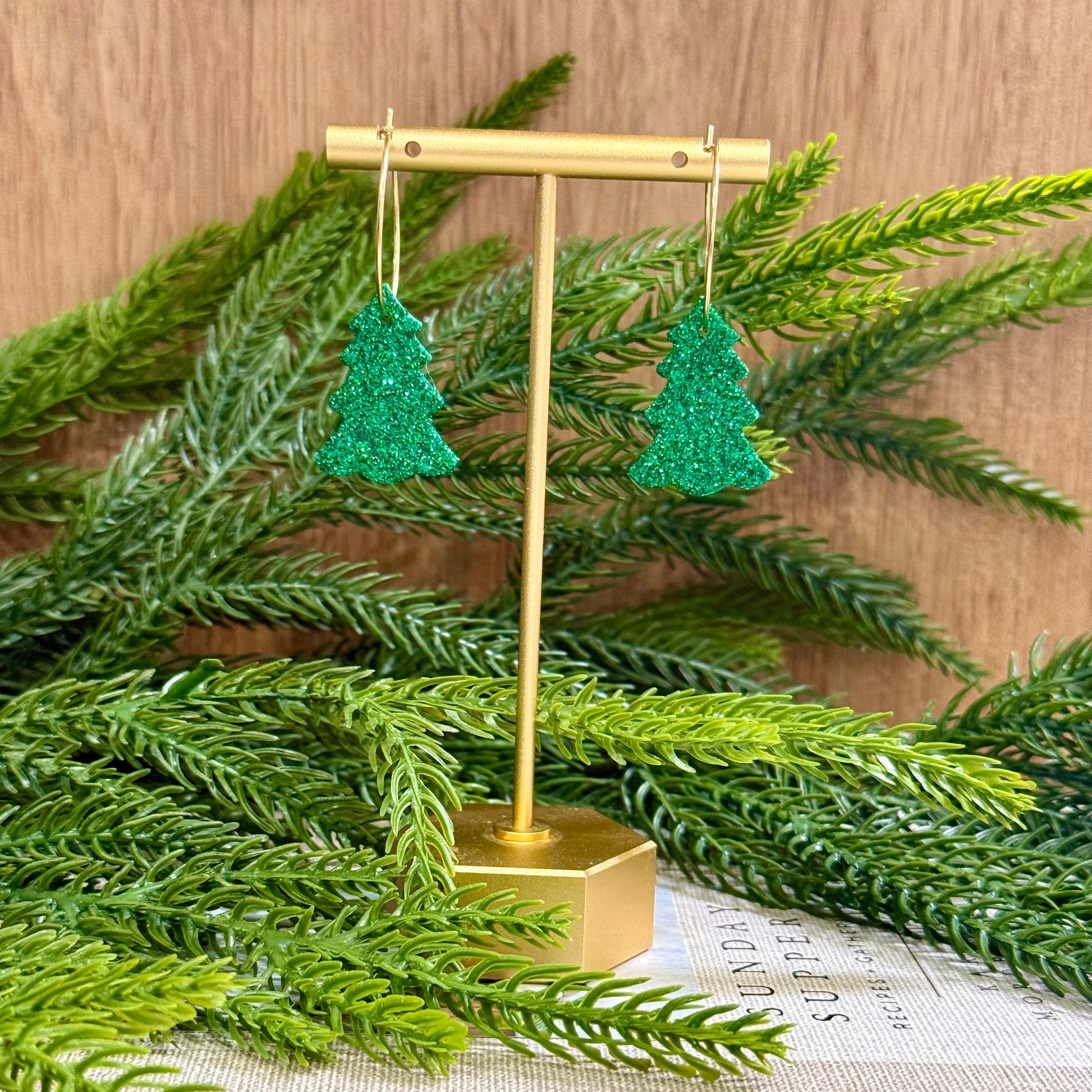 Green glittery tree-shaped earrings on a gold stand with green foliage in the background.
