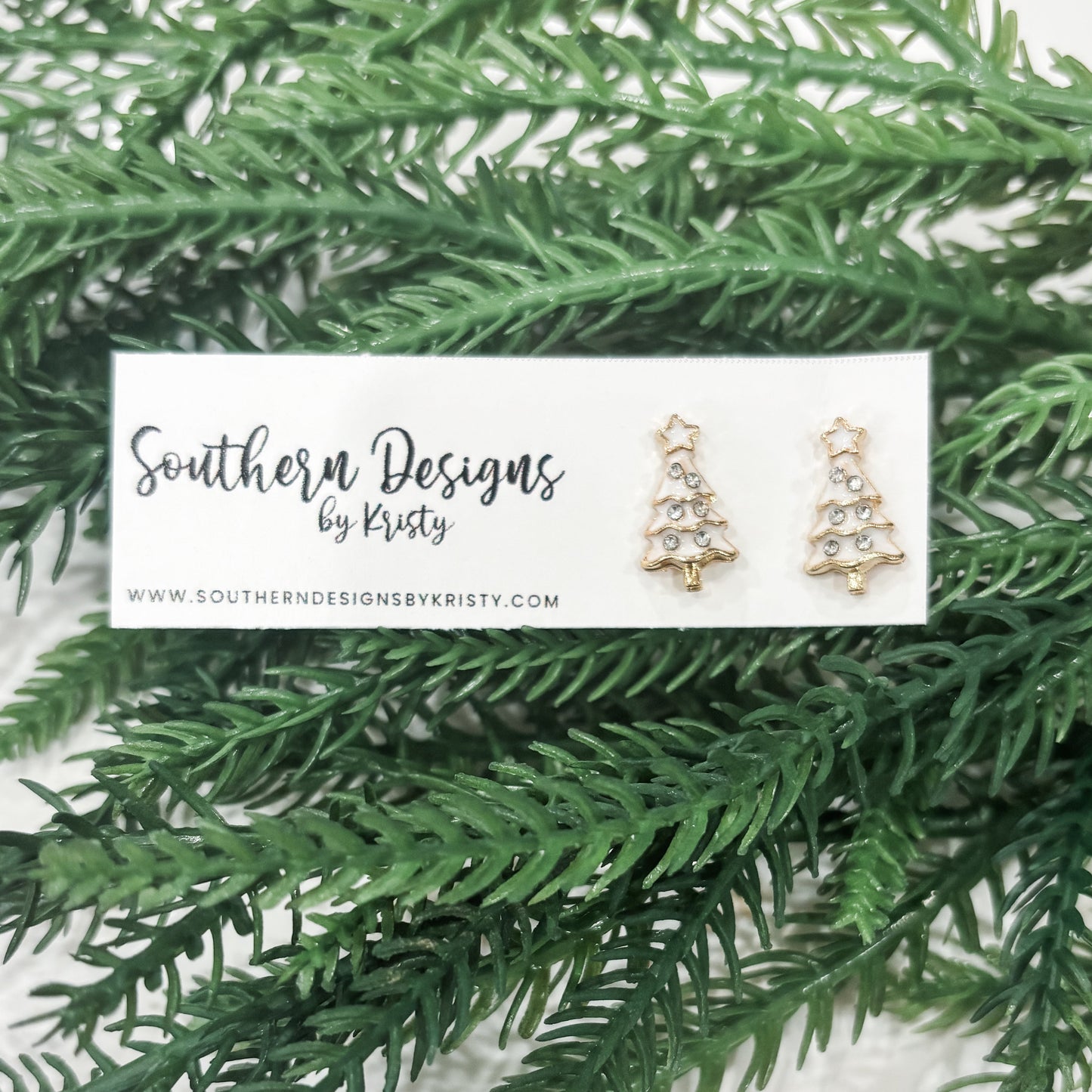 A pair of Christmas tree-shaped stud earrings displayed on a card with 'Southern Designs by Kristy' written on it, placed on a green pine needle background.