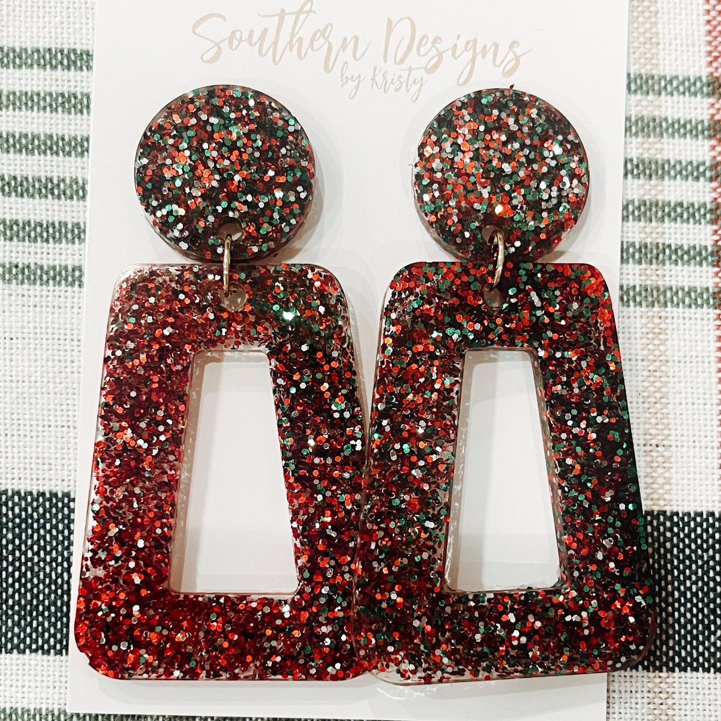 A pair of glittery red and black Christmas-themed dangle earrings displayed on a textured fabric background with 'Southern Designs' branding.