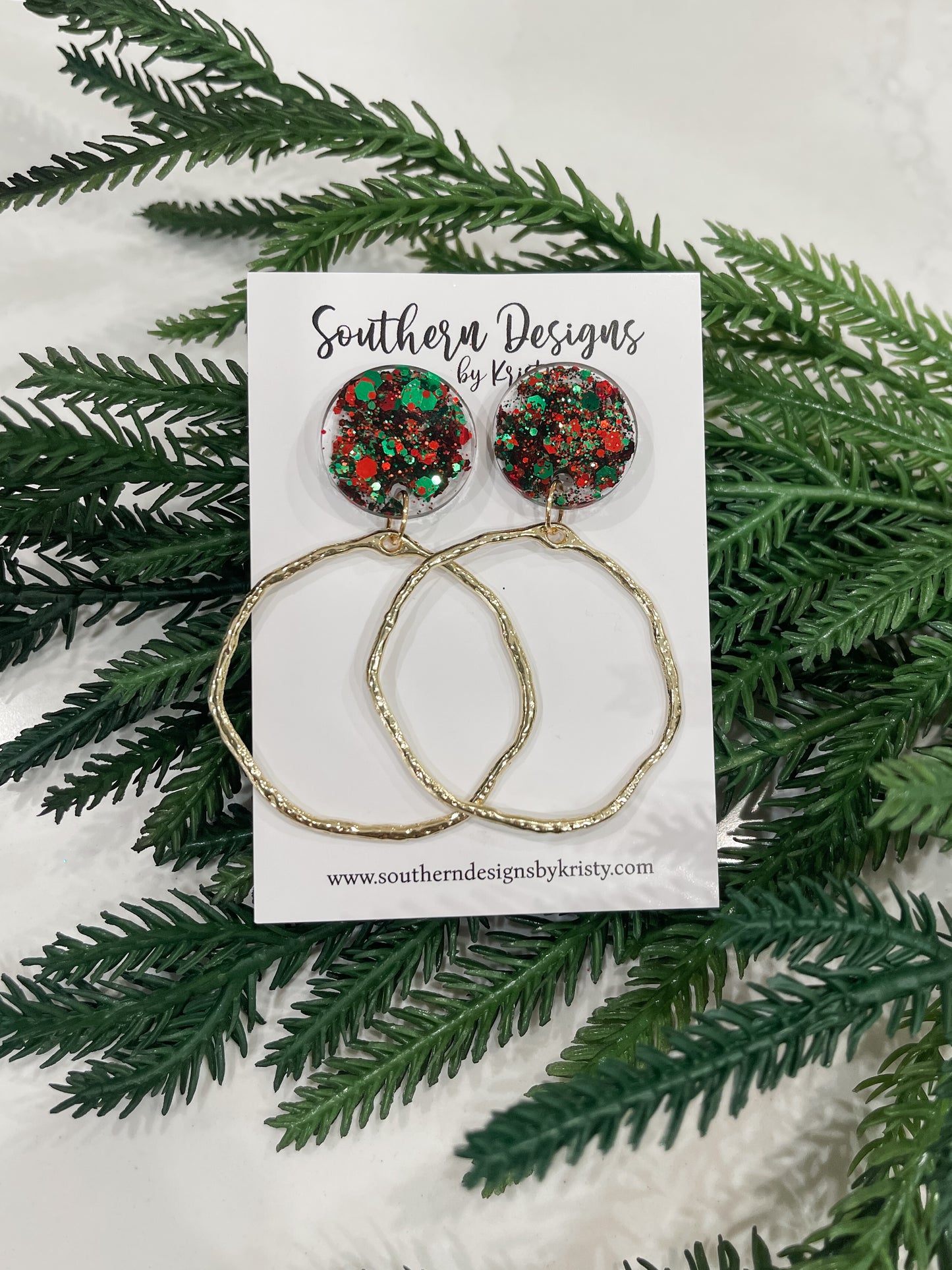 A pair of dangle earrings with red and green circular designs, displayed on a white card with 'Southern Designs by Kristy' printed on it, surrounded by green pine branches.