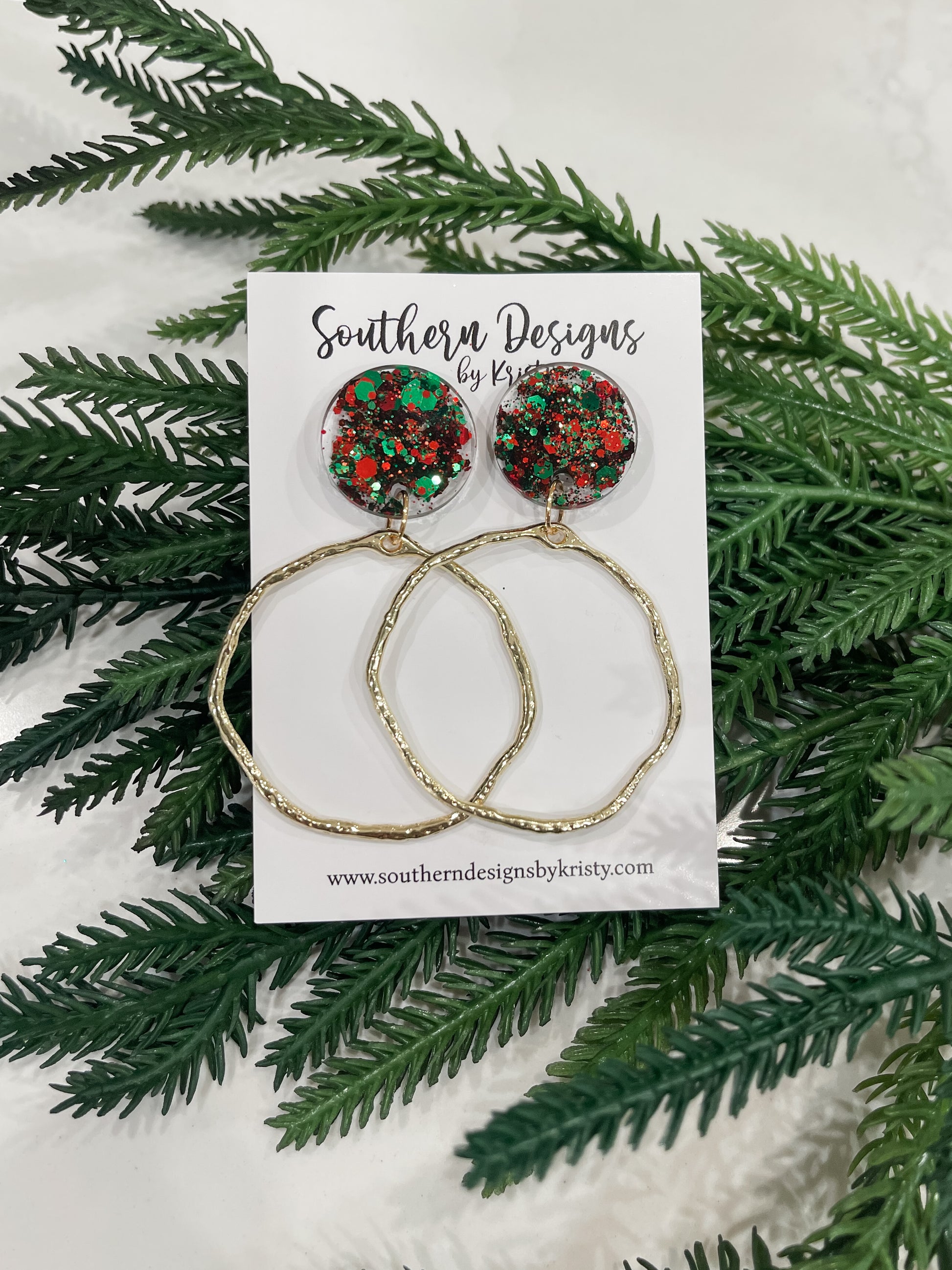 A pair of dangle earrings with red and green circular designs, displayed on a white card with 'Southern Designs by Kristy' printed on it, surrounded by green pine branches.
