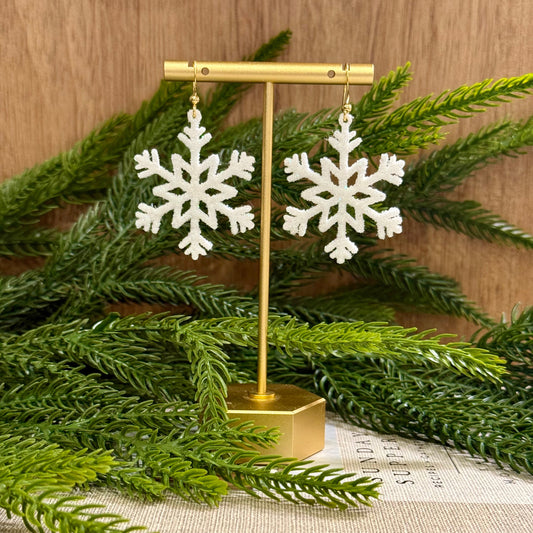 glitter Snowflake earrings on a gold stand with greenery in the background