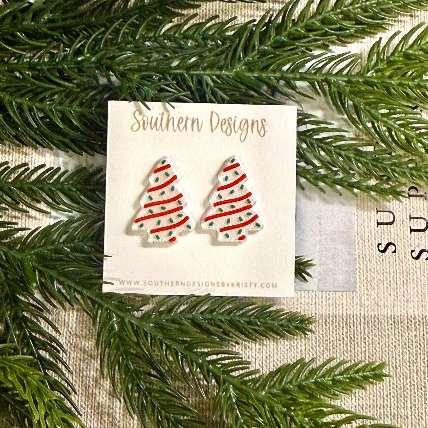 Pair of Christmas tree earrings on a card with 'Southern Designs' branding, surrounded by green foliage.