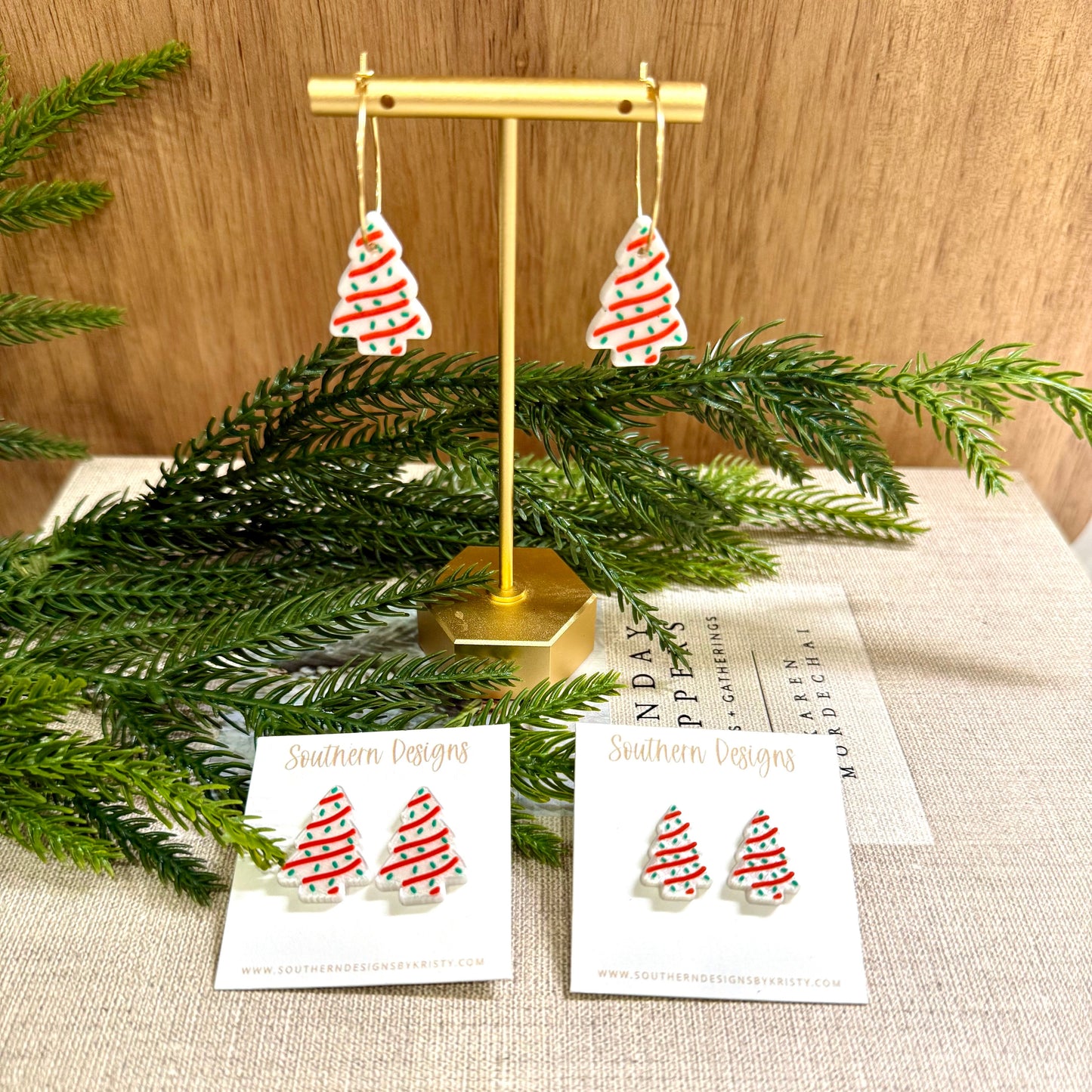 Christmas tree earrings on a stand with greenery and branded cards on a textured surface.